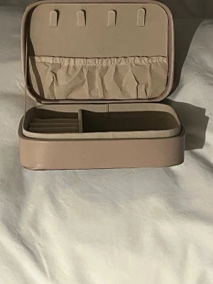 Beige smyckesväska i skinnimitation - Snygg och stilren beige smyckesväska i skinnimitation med flera fack och elastisk ficka på insidan. Perfekt för att organisera ringar, örhängen och halsband när du är på språng. Väskan har en mjuk, rektangulär form och stängs med dragkedja.