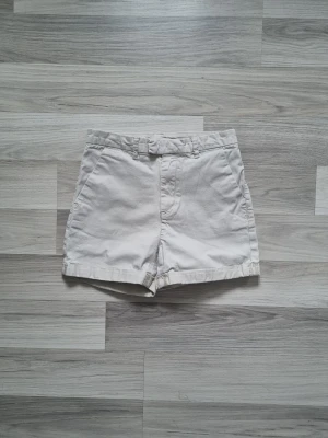Beige shorts från H&M, strl 146 - Snygga beige shorts från H&M i klassisk modell med uppvikta benslut och bälteshällor. Shortsen har två sidofickor fram och två bakfickor med knappar. Tillverkade i mjuk bomull, perfekta för varma dagar.