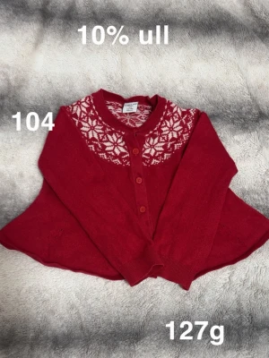 Röd kofta med snöflingemönster - Mysig röd kofta från Polarn O. Pyret med vita snöflingor runt halsen. Tröjan har långa ärmar, rund halsringning och knäppning framtill med matchande röda knappar. Innehåller 10% ull för extra värme.🌸 Fint använt skick.. Storlek 104