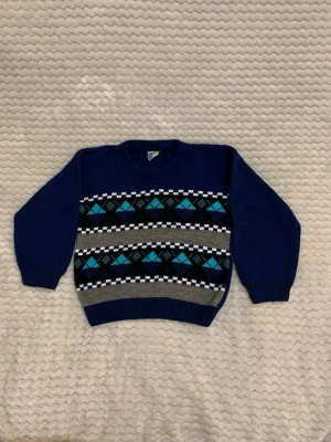 Blå stickad tröja med mönster - Mysig blå stickad tröja från Cool Club med klassiskt argyle-mönster i blått, turkos, svart och vitt. Tröjan har långa ärmar, rund hals och ribbade muddar. Perfekt för kyliga dagar och snygg till jeans eller chinos.