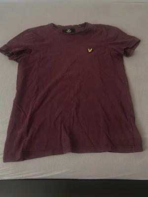 Vinröd t-shirt från Lyle & Scott - Säljer en vinröd t-shirt från Lyle & Scott med klassisk rund hals och det ikoniska guldiga logomärket broderat på bröstet. T-shirten är i mjuk bomull och har en enkel, stilren design med korta ärmar.