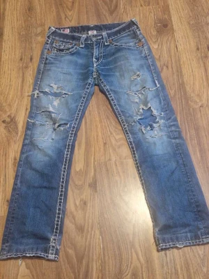 True Religion Ricky Super T blå jeans - Säljer ett par blå True Religion Ricky Super T jeans med slitna detaljer och stora hål framtill. Klassiska kontrastsömmar och ikoniska bakfickor med vita broderier och knappar. Raka ben och låg midja. Perfekta för dig som gillar en edgy streetstil. 