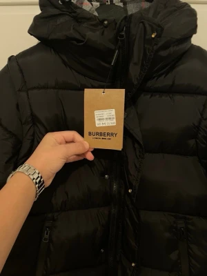 Svart dunväst från Burberry - Säljer en svart dunväst från Burberry med huva och dragkedja framtill. Västen har två fickor med dragkedja och klassiskt rutigt foder i huvan. Perfekt för lager på lager och riktigt snygg till höst- och vinterlooken.