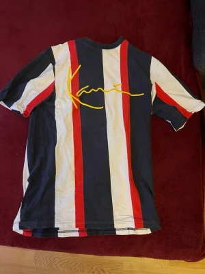 Randig t-shirt från Karl Kani - Säljer en kortärmad t-shirt från Karl Kani med breda vertikala ränder i svart, vitt och rött. Stor gul broderad logga på bröstet ger en unik touch. Tillverkad i mjuk bomull, perfekt för en avslappnad streetwear-look.