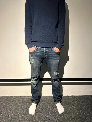 Jack & Jones jeans med slitningar - Säljer ett par blå jeansbyxor från j&j med slitningar och detaljerade sömmar på bakfickorna. Modellen har normal passform och klassisk femficksdesign. Jeansen har en något tvättad look och är tillverkade i tåligt denimtyg.