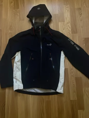 Marinblå och vit vindjacka från Pelle P - Säljer en marinblå och vit vindjacka från Pelle P med huva och dragkedja framtill. Jackan har vita sidopaneler, logga på bröstet och ärmen samt coola detaljer som röda snören och tryckta koordinater på ena ärmen. Perfekt för dig som gillar sportig stil.