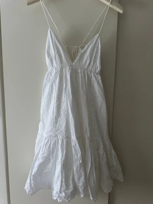 Vit sommarklänning från Zara - Supersöt vit kortklänning från Zara med tunna axelband, broderade blommönster och volangkant. Klänningen har v-ringning, öppen rygg med knyt och är tillverkad i bomull. 