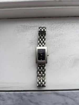 Gucci klocka  - Stilren klocka från Gucci i silverfärgad metall med rektangulärt svart ur och länkarmband med ovala länkar. Klassisk design med diskret logga på urtavlan och graverad baksida. Perfekt accessoar för dig som gillar exklusiva detaljer.OBS VET EJ OM DEN FUNKAR!!!!!! kan inget om klockor kom med bud