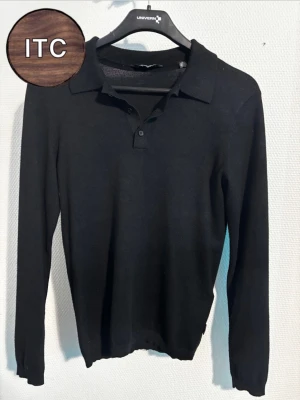 Svart pikétröja  - Klassisk svart pikétröja från ITC i mjuk ull. Tröjan har lång ärm, krage och knäppning framtill med två knappar. Perfekt för dig som gillar stilrena och tidlösa plagg. Passar lika bra till jeans som till chinos.