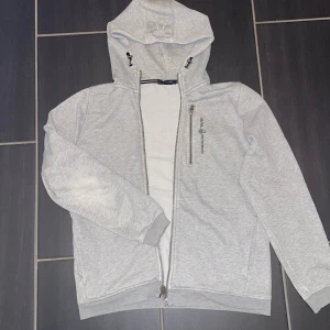 Ljusgrå hoodiejacka från Sail Racing - Ljusgrå hoodiejacka från Sail Racing med dragkedja framtill och bröstficka med logga. Jackan har huva med snörning och ribbade muddar. Perfekt för chill dagar och enkel att matcha med jeans eller joggers.