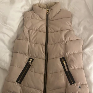 Beige dunväst från Joules - Snygg beige dunväst från Joules med quiltad design och svarta detaljer vid dragkedjorna. Västen har hög krage och två fickor framtill med dragkedja. Insidan är fodrad med blått och rött rutigt tyg. Perfekt för lager-på-lager-stil. Nypris ca 1000-2000