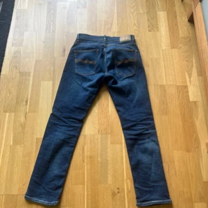  Grim Tim Nudie Jeans Indigo Myth, W31 L30 - Snygga mörkblå jeans från Nudie Jeans med klassisk femficksdesign och kontrastsömmar. Modellen har rak passform och normal midja, tillverkade i slitstarkt bomullsjeansmaterial. Perfekt för dig som gillar stilrena och tidlösa jeans.