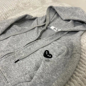 Grå hoodie från Comme des Garçons Play - Grå hoodie från Comme des Garçons Play med dragkedja och huva. Ikoniskt svart hjärta med ögon på bröstet och stort hjärta på ryggen. Mjuk bomullskvalitet, ribbade muddar och snörning i huvan. Perfekt för en chill och stilren look.