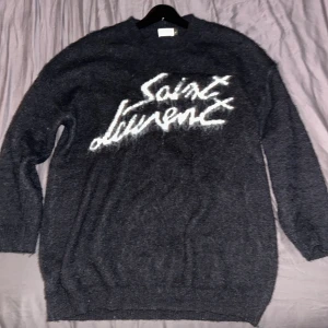 Svart stickad Saint Laurent tröja M - Svart stickad tröja från Saint Laurent med vit logga i graffiti-stil över bröstet. Tröjan har rund hals, ribbade muddar och lång ärm. Mjuk och lite luddig känsla, perfekt för dig som gillar exklusiva streetwear-vibbar.