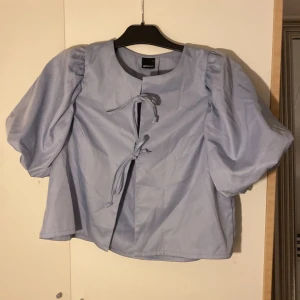Ljusblå blus med puffärm Gina Tricot - Supersöt ljusblå blus från Gina Tricot i storlek S. Blusen har puffiga korta ärmar och två knytband framtill som ger en unik look. Tillverkad i mjuk bomull med rund hals och loose passform. Perfekt för dig som gillar trendiga detaljer och stilren design.