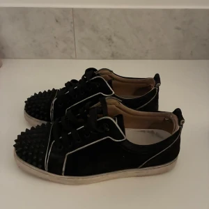 Svarta sneakers med nitar från Louboutin - Unika svarta sneakers från Christian Louboutin med coola svarta nitar på tån, vita kontrastsömmar och den klassiska röda sulan. Skorna har snörning och är tillverkade i mocka med detaljer i skinn. Perfekt för dig som vill sticka ut med din stil. 2 spikar saknar, inga andra defekter skick 7/10 storlek 40,5 (passar 42)