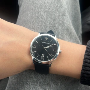 Emporio Armani svart klocka i läder - Stilren klocka från Emporio Armani med svart urtavla, romerska siffror och datumvisning. Boetten är i silverfärgad metall och armbandet är gjort av svart läder med klassiskt spänne. Perfekt accessoar för en snygg och tidlös look. Knappt använd, köpt för 2000