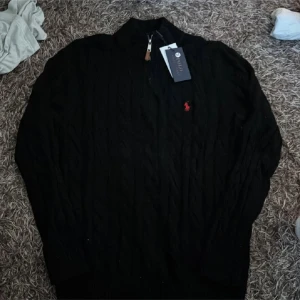 Svart stickad tröja från Ralph Lauren - Svart stickad tröja från Ralph Lauren med klassisk kabelstickning och röd broderad logga på bröstet. Tröjan har lång ärm, hög krage och half zip-dragkedja med läderdetalj. Perfekt för dig som gillar stilren och tidlös design.