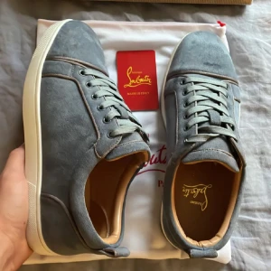 Christian Louboutin skor, storlek 41-42 - Snygga sneakers från Christian Louboutin i grå mocka med klassisk röd sula. Skorna har rund tå, platt vit sula och grå snörning. Insidan är fodrad i beige skinn för extra komfort. Perfekta för dig som vill ha en stilren och lyxig look.