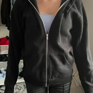 Svart hoodie med dragkedja och huva - Svart hoodie med hel dragkedja framtill och stor huva. Tröjan har långa ärmar, ribbade muddar och fickor på magen. Tillverkad i mjuk bomull, perfekt för chill dagar. Enkel och stilren design som funkar till allt.