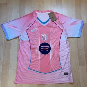 Barça Lamine Yamal 10 rosa Nike tröja - Snygg FC Barcelona fotbollströja från Nike i rosa och ljusblå toner med tryck Lamine Yamal 10 på ryggen. Tröjan har korta ärmar, V-ringad krage med blå kant och detaljerade linjemönster framtill. Materialet är lätt och andas, perfekt för match eller träning.