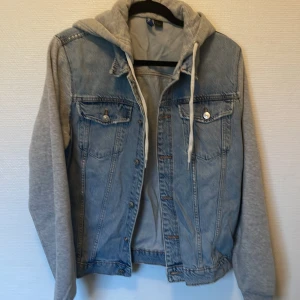 Jeansjacka med grå hoodie från Divided - Snygg jeansjacka från Divided med ljusblå denim och grå sweatshirt-ärmar samt huva. Jackan har knappar framtill, bröstfickor och dragsko i huvan. Perfekt för en chill streetstyle-look.