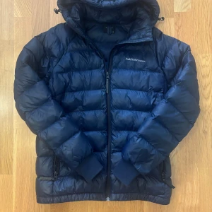 Mörkblå dunjacka Peak Performance 160 - Snygg mörkblå dunjacka från Peak Performance med huva och dragkedja framtill. Jackan har quiltad design, ribbade muddar och två fickor med dragkedja. Perfekt för kalla dagar och riktigt skön att bära.