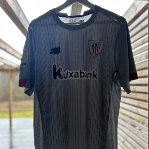 Athletic Bilbao bortatröja New Balance XL - Snygg Athletic Club Bilbao bortatröja från New Balance i ljusgrått med tunna svarta ränder. Kortärmad modell med röda detaljer på ärmsluten och La Liga-märke på ena ärmen. Tillverkad i lätt och ventilerande polyester, perfekt för match eller träning men även en tröja att gå runt med i vardagen. Tröjan är äkta och köpt från TK Maxx, 9/10 skick nästan aldrig använt då jag inte hejar på de den har bara hängt i min garderob.