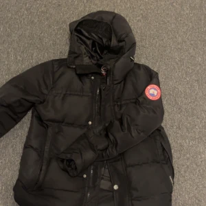 Svart dunjacka från Canada Goose - Säljer en svart dunjacka från Canada Goose med huva och klassisk logga på ärmen. Jackan har dragkedja framtill, knappar och två sidofickor. Perfekt för kalla dagar och riktigt snygg streetstil.