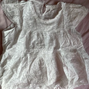 Vit broderad blus från Liance - Supersöt vit blus från Liance med broderade blommor och spetsdetaljer vid halsringning och ärmslut. Blusen har korta ärmar och en luftig, volangig passform. Perfekt för sommarens alla tillfällen när du vill känna dig extra fin.