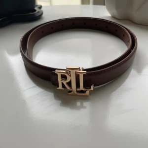 Brunt skärp från Ralph Lauren RL - Snyggt brunt bälte från Ralph Lauren med ett stilrent RL-spänne i guldmetall. Bältet är gjort i slätt skinn och har klassisk design med flera hål för justerbar passform. Perfekt accessoar för att lyfta din outfit med en lyxig touch.