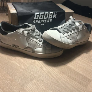 Golden Goose Superstar sneakers vit/grå - Golden Goose Superstar sneakers i vitt skinn med grå mockastjärna på sidan och grå snörning. Snygg kontrast med svart häl och platt sula. Ikonisk streetstil med GGDB-detaljer och box medföljer. Perfekt för dig som gillar exklusiva sneakers med unik design. Bara en liten defekt på sulorna, se bild 5, men enkelt att fixa 