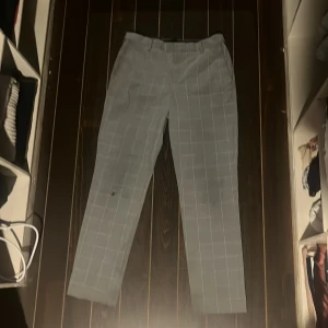 Grå rutiga kostymbyxor från H&M - Grå kostymbyxor från H&M med vitt rutmönster. Klassisk rak passform med bälteshällor och bakficka med knapp. Perfekta för en clean och stilren look. Materialet är mjukt och lätt, vilket gör dem bekväma hela dagen.