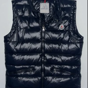 Svart dunväst från Moncler - Snygg svart dunväst från Moncler med glansig finish och klassisk quiltad design. Västen har dragkedja framtill, två snedställda fickor med dragkedja och Moncler-logga på bröstet. Perfekt för lager-på-lager och streetstyle.