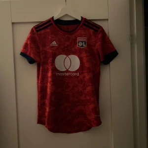 Röd Olympique Lyonnais fotbollströja  - Säljer en röd Olympique Lyonnais fotbollströja från Adidas i storlek S. Tröjan har mörkblå detaljer, korta ärmar och ett camouflagemönster. Tillverkad i lätt AEROREADY-material, perfekt för träning, match eller att gå runt med i vardagen. Mastercard-logga på bröstet. Helt äkta köptes på TK Maxx och är i princip aldrig använd då jag ej hejar på de.