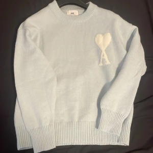Ljusblå stickad tröja från Ami - Snygg ljusblå stickad tröja från Ami med ett vitt hjärta och bokstaven A broderat på bröstet. Tröjan har rund hals, ribbade muddar och är långärmad. Perfekt för dig som gillar stilrena detaljer och mjuka material.