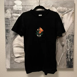 Svart t-shirt med nalletryck Hendrix - Svart t-shirt från Hendrix med ett litet tryck av en orange nalle och gröna blad på bröstet. Klassisk passform med rund hals och korta ärmar. Perfekt för dig som gillar unika prints och vill sticka ut lite extra.