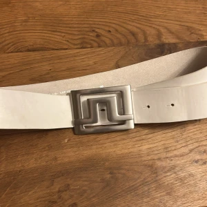 J Lindberg bälte - Snyggt vitt bälte i äkta skinn, tillverkat i Italien. Bältet har ett brett band och ett unikt, geometriskt spänne i silverfärg som verkligen sticker ut. Perfekt accessoar för att lyfta din outfit med en modern touch.