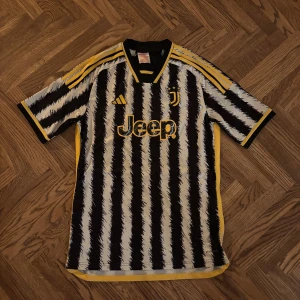 Juventus fotbollströja Adidas 15-16 år - Juventus fotbollströja från Adidas med svart och vit randigt mönster och gula detaljer. Tröjan har klubbmärke med tre stjärnor, Jeep-logga på bröstet och klassiska Adidas-ränder på axlarna.