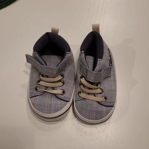 Grå sneakers från Newbie, strl 19/20 - Söta grå sneakers från Newbie i textil med beige snören och kardborreband. Skorna har vit sula med svarta detaljer och prickigt mönster undertill för extra grepp. Broderad Newbie-logga på sidan och smidig ögla bak för enkel påtagning.