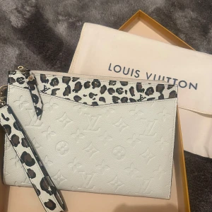 Vit clutch med leoparddetaljer NYSKICK - Snygg clutch i vit skinn med coola leoparddetaljer på dragkedja och överkant. Väskan har en avtagbar handledsrem och guldiga metalldetaljer. Perfekt för att lyfta din outfit med lite extra edge.