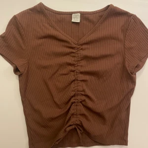 Brun ribbad topp med snörning från Lindex - Säljer en brun ribbad topp från Lindex med korta ärmar och v-ringning. Toppen har snygg snörning framtill som ger en cool rynkad effekt och är croppad i modellen. Perfekt för dig som gillar enkla men trendiga detaljer.