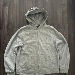 Grå hoodie från Polo Ralph Lauren - Klassisk grå hoodie från Polo Ralph Lauren med dragkedja framtill, huva och känguruficka. Broderad marinblå logga på bröstet. Mjuk insida och avslappnad passform, perfekt för chill dagar. Passar som S.