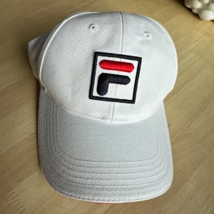 Vit FILA keps med broderad logga - Snygg vit keps från FILA med broderad logga i svart och rött framtill. Kepsen har böjd skärm och justerbar rem baktill. Insidan av skärmen är röd för extra kontrast. Tillverkad i 100% bomull. Kepsen är i nyskick! 