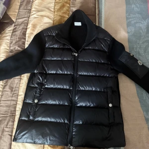 Svart Moncler pufferjacka med stickade ärmar - Snygg svart pufferjacka från Moncler med glansig dunfylld framsida och ribbstickade ärmar och rygg. Jackan har dragkedja framtill, två fickor med tryckknappar och Moncler-logga på ärmen. Perfekt för kyliga dagar och ger en riktigt clean look. Kan sänka priset vid snabba köp