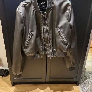 Svart bomberjacka i skinnimitation Zara XS - Snygg svart bomberjacka från Zara i skinnimitation med silvriga dragkedjor och ribbade muddar. Jackan har två snedställda fickor med dragkedja och klassisk bomberkrage. Perfekt för dig som vill ha en stilren och trendig look.