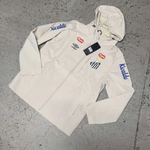 Beige vindjacka från Umbro Santos FC - Snygg beige vindjacka från Umbro med Santos FC-logga på bröstet och blåa Kicaldo-tryck på ärmarna. Jackan har huva, hel dragkedja och sportiga detaljer. Perfekt för dig som gillar fotboll och vill ha en stilren look.
