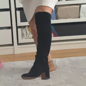 Svarta overknee mockastövlar med klack - Säljer ett par svarta overknee stövlar i mjuk mocka med rund tå och stabil blockklack på ca 6 cm. Stövlarna har en stilren och slimmad siluett som går högt upp över knät. Perfekta för dig som vill ha en edgy look till hösten och vintern.
