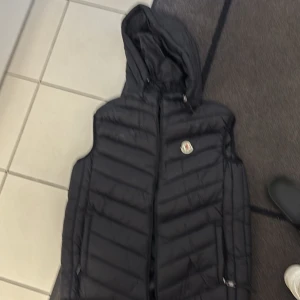 Svart dunväst med huva från Moncler - Svart dunväst från Moncler med huva och klassisk logga på bröstet. Västen är quiltad och har dragkedja framtill. Tillverkad i glansig polyester och fylld med 90% gåsdun. Perfekt för lager-på-lager och snygg streetstil.