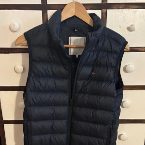 Mörkblå dunväst från Tommy Hilfiger - Snygg mörkblå dunväst från Tommy Hilfiger med klassisk quiltad design och diskret logga på bröstet. Västen har hög krage och dragkedja framtill. Perfekt att slänga över en hoodie för extra värme under kyliga dagar. Storlek 164 som passar ungefär som XS. Skriv vid frågor, byten kan även vara aktuellt.